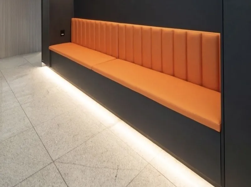 Parede revestida em ACM preto com banco laranja estofado e iluminação em LED